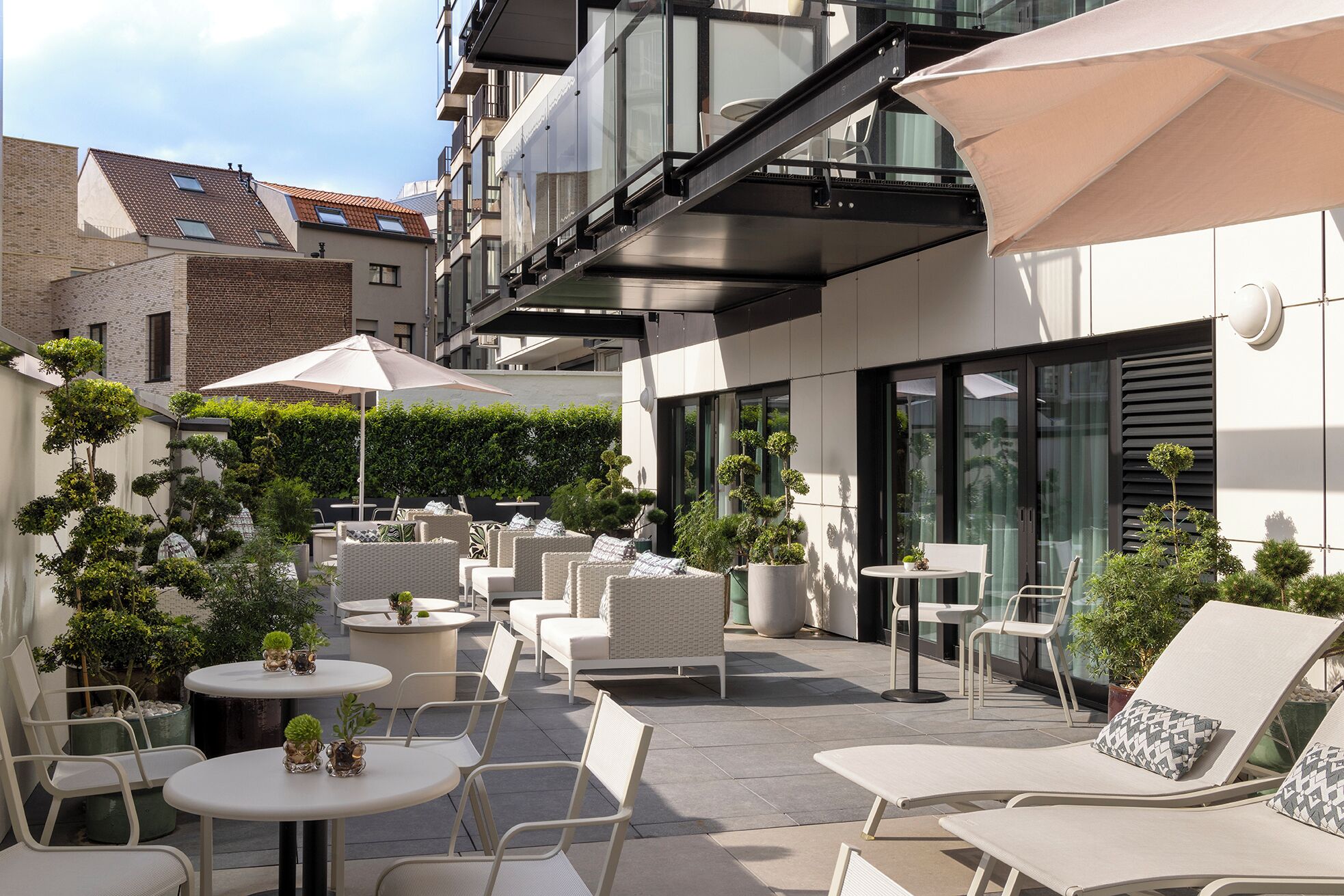 Un patio en la azotea con mobiliario exterior, que incluye sillas y mesas, rodeado de vegetación y edificios modernos.