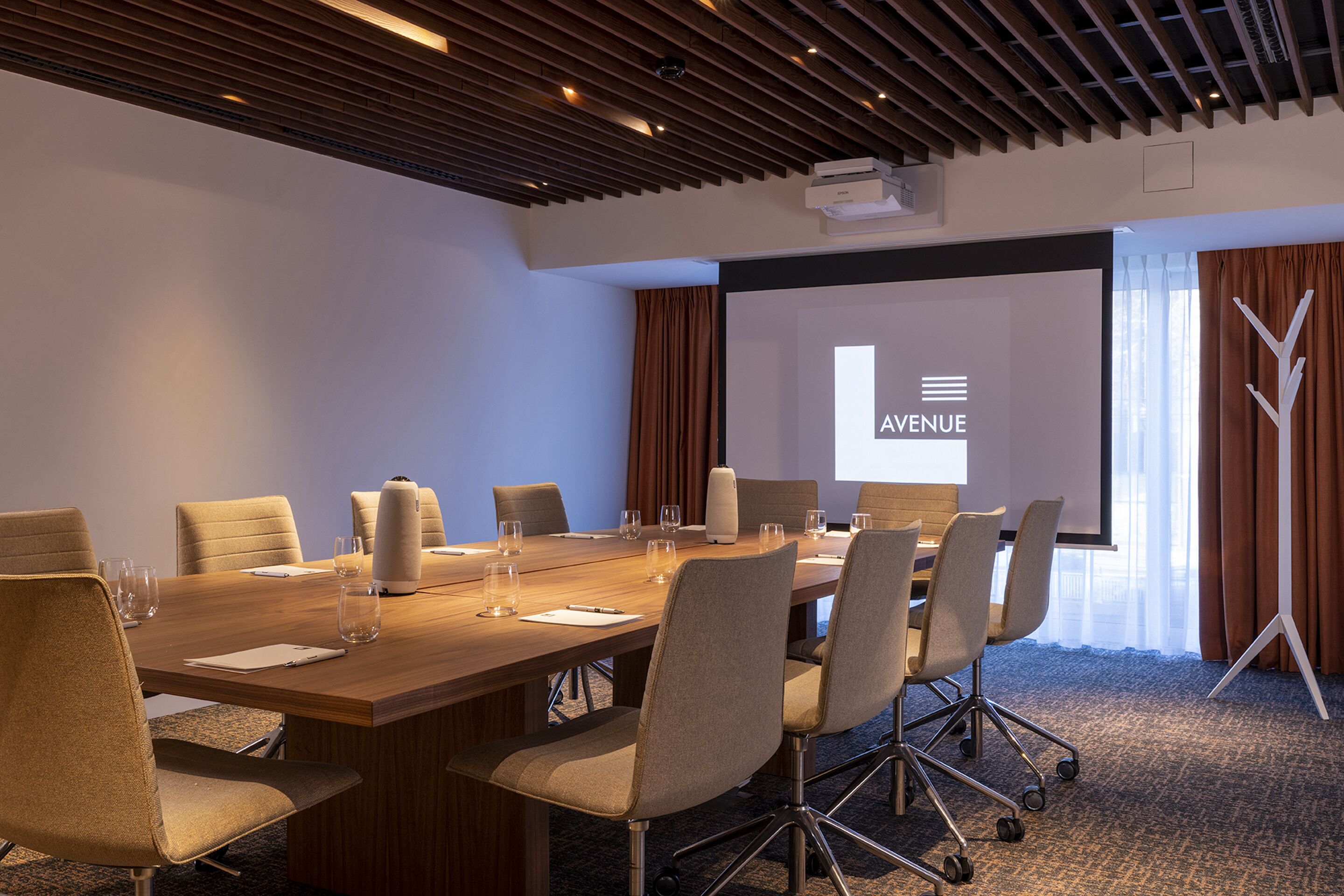 Uma sala de conferências com uma mesa comprida, várias cadeiras, uma tela de projetor e uma janela grande com cortinas.