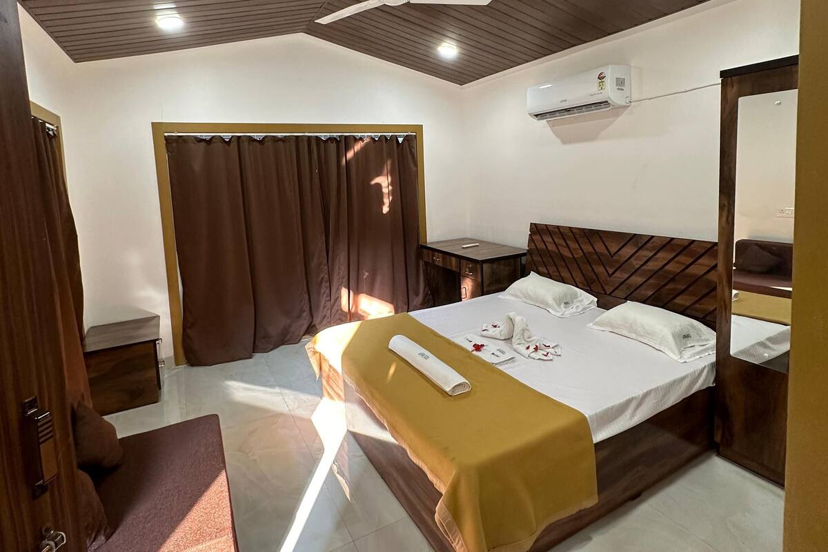 A hotel room with a bed, a ceiling fan, an air conditioning unit, a bedside table, and a mirror.