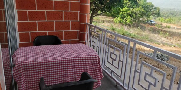 A balcony with a checkered tablecloth, a chair, and a view of a rural landscape with trees and a dirt path.