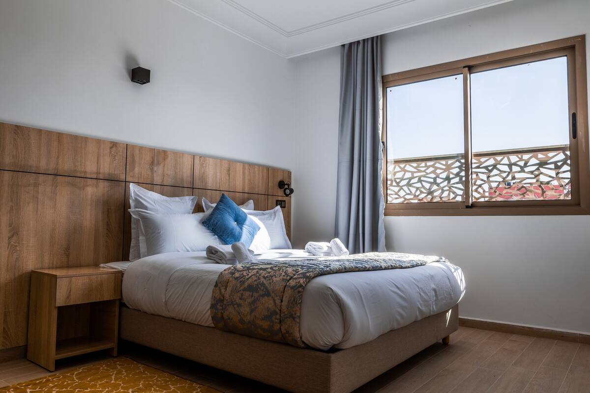 A bedroom with a wooden headboard, a large window with a view, and a neatly made bed with pillows and a blanket.