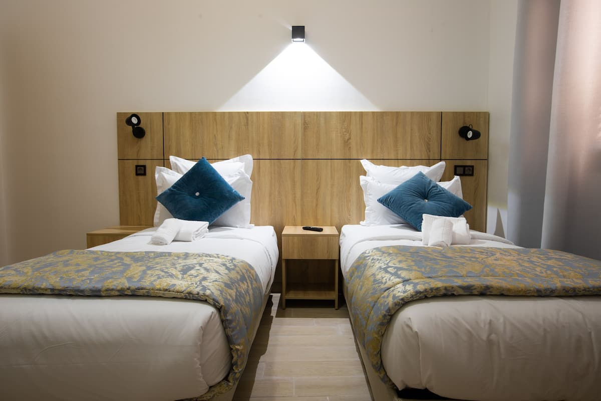 A hotel room with two beds, each with a wooden headboard, white bedding, and blue pillows. There is a nightstand between the beds with a remote control on it.