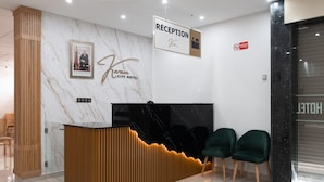 Ein Hotel-Empfangsbereich mit Marmorwand, einer Rezeption mit schwarzer Arbeitsplatte und zwei grünen Sesseln.