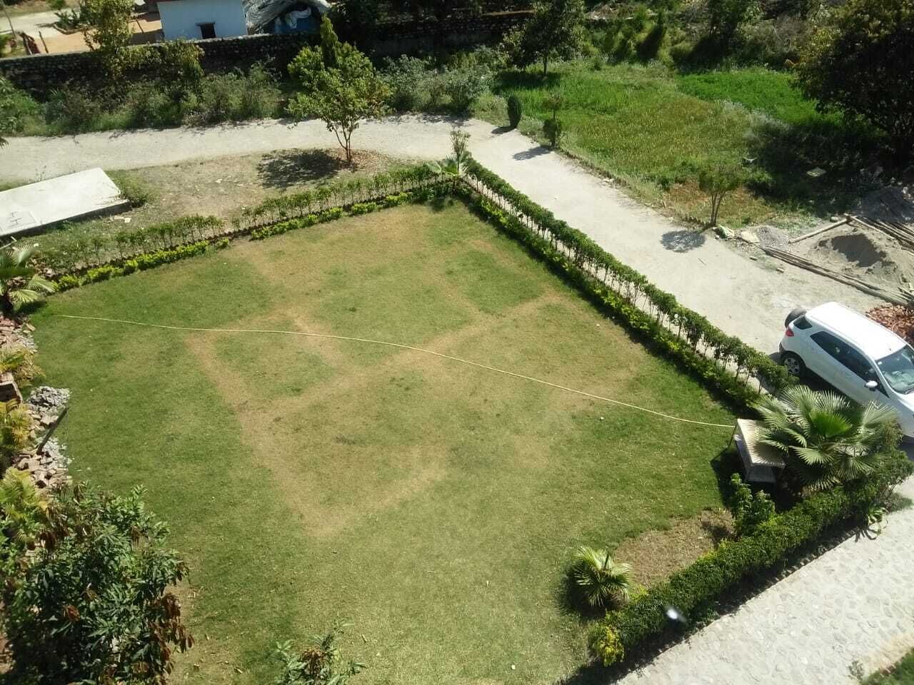 A square lawn with a paved border, surrounded by greenery and a parked car.