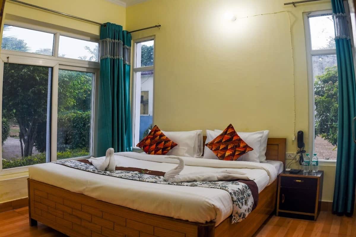 Una cama bien hecha con un cabecero de madera, dos almohadas con un estampado geométrico y una almohada con forma de cisne. Hay una mesita de noche pequeña con un teléfono y una botella. La habitación tiene ventanas grandes con cortinas, y las paredes están pintadas de amarillo.
