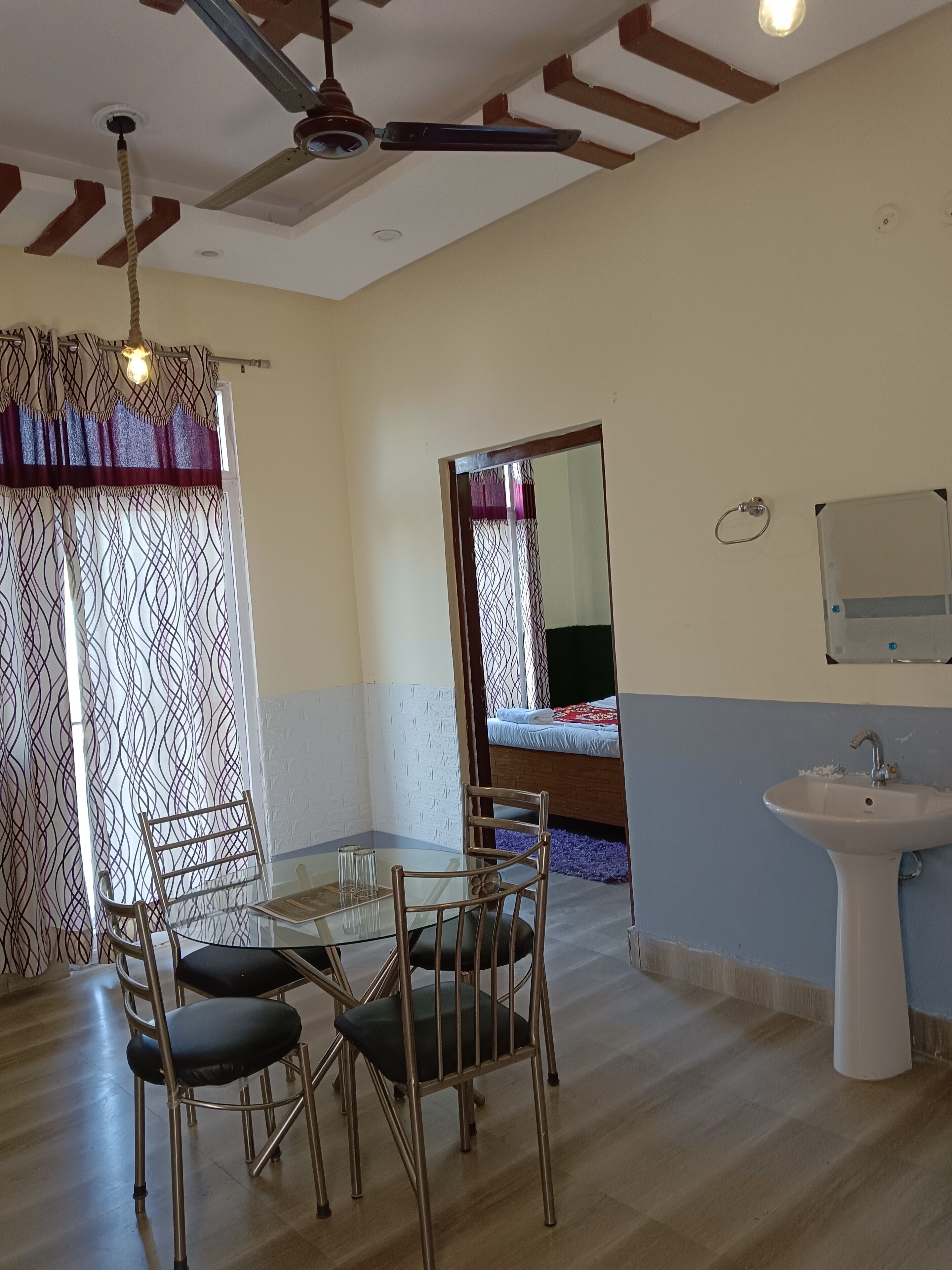 A room with a dining table and chairs, a ceiling fan, a bed with a patterned bedspread, and a sink.