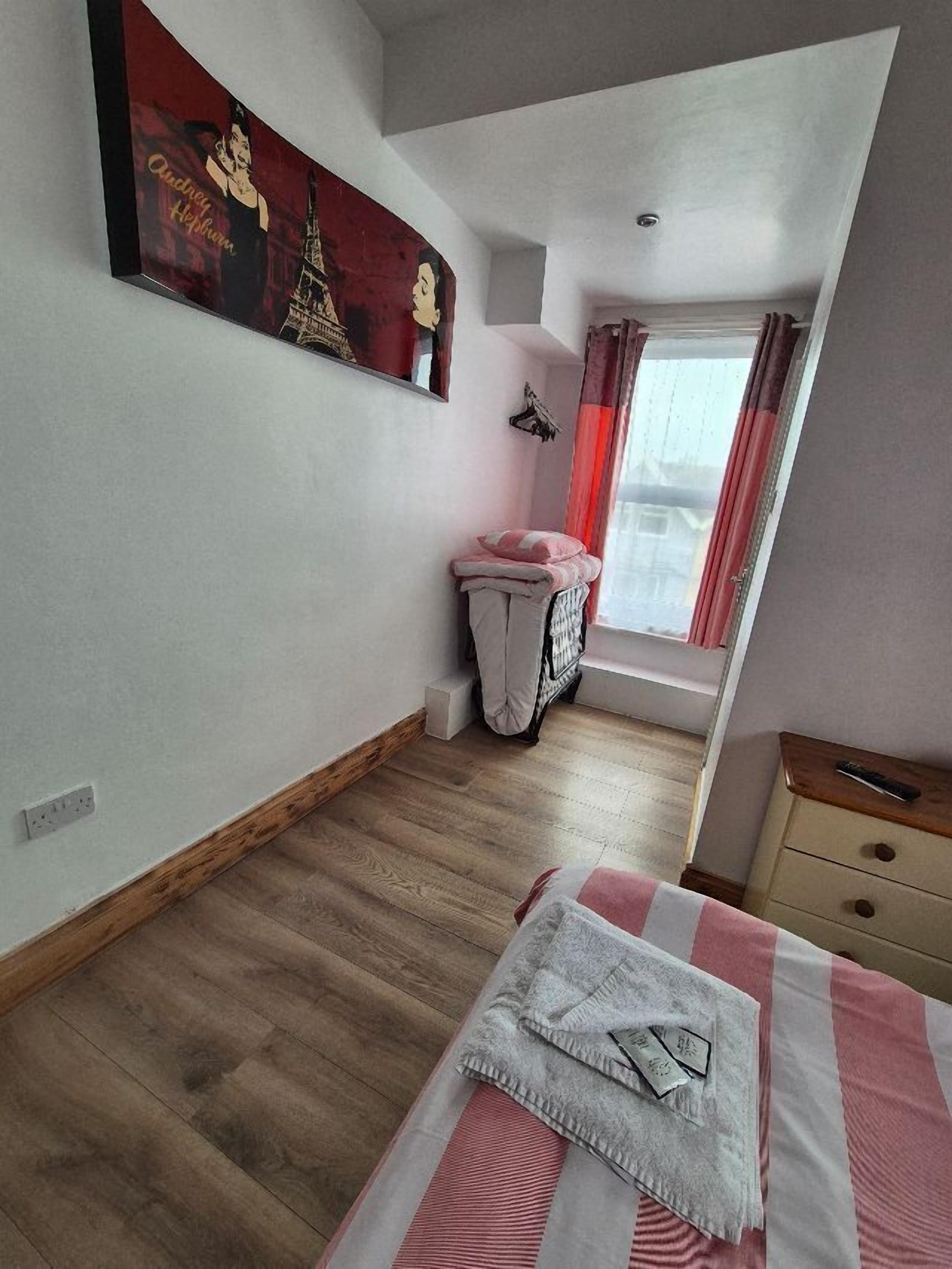 Un dormitorio con suelo de madera, una cama con manta a rayas, una mesita de noche, una ventana con cortinas y un cuadro en la pared.