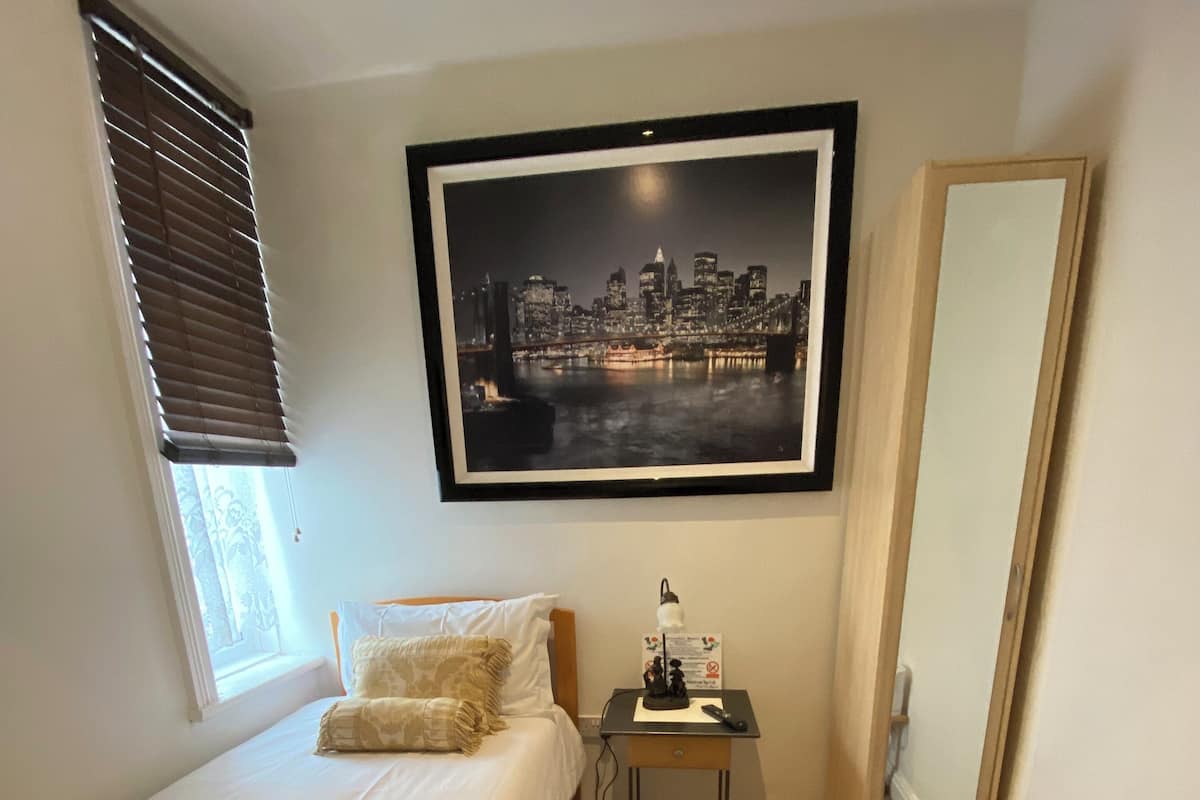 Quarto com cama, mesa de cabeceira, quadro com vista urbana emoldurada, janela com persianas e piso de madeira.