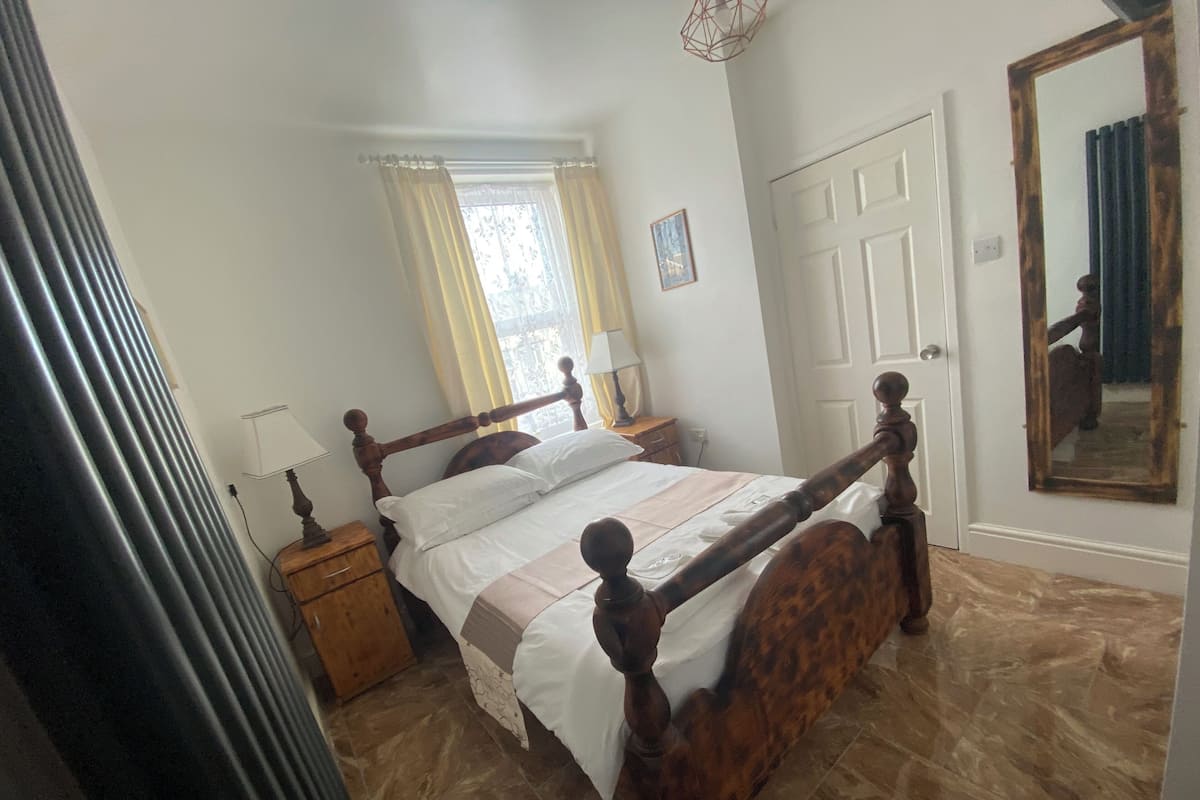 Quarto com cama de dossel de madeira, mesa de cabeceira, luminária, janela com cortinas, porta e espelho.