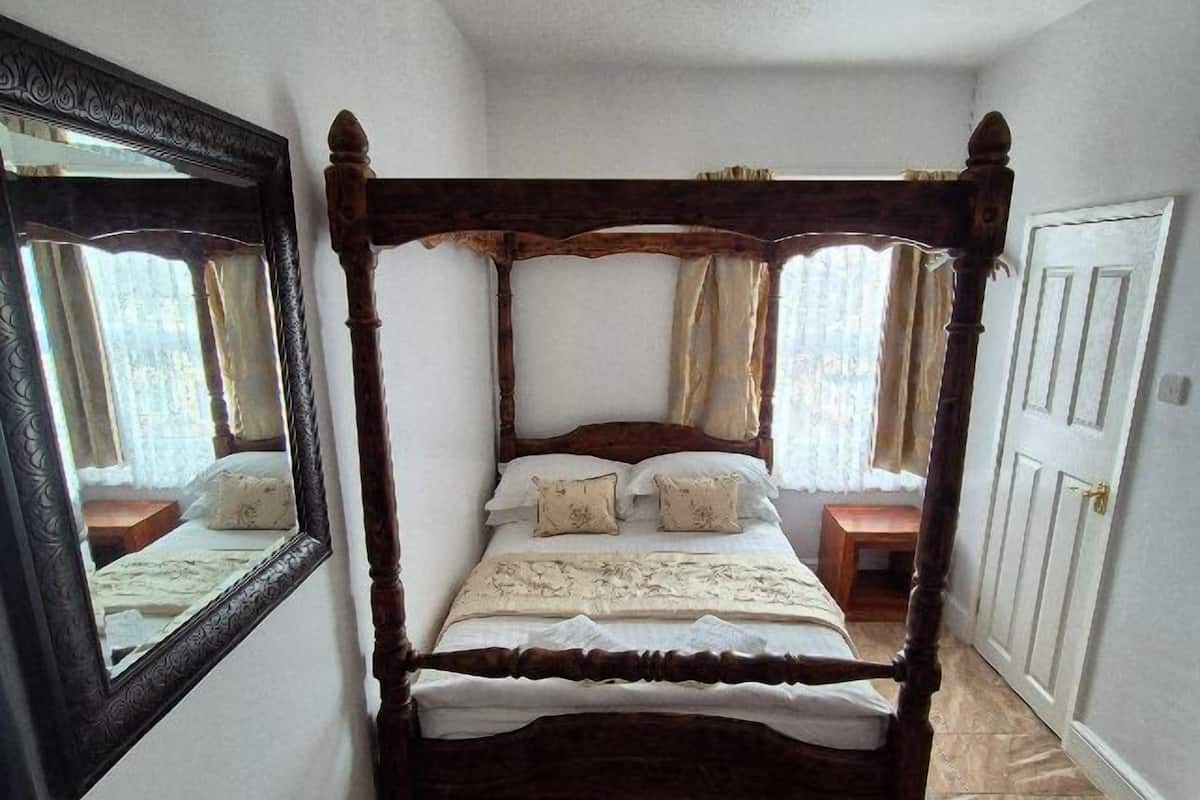 Uma cama com dossel e estrutura de madeira, um espelho à esquerda e uma porta à direita.