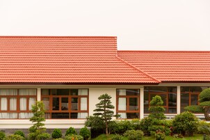 Un edificio con un tetto in tegole rosse, numerose finestre con tende e un giardino curato con diverse piante e arbusti.