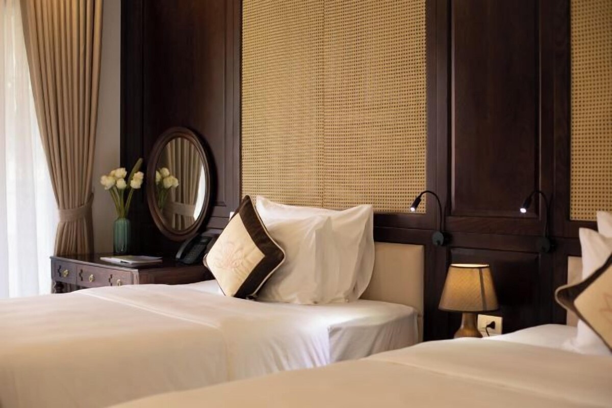Habitación de hotel con dos camas, una mesita de noche con lámpara, un espejo y una ventana con persiana tejida.