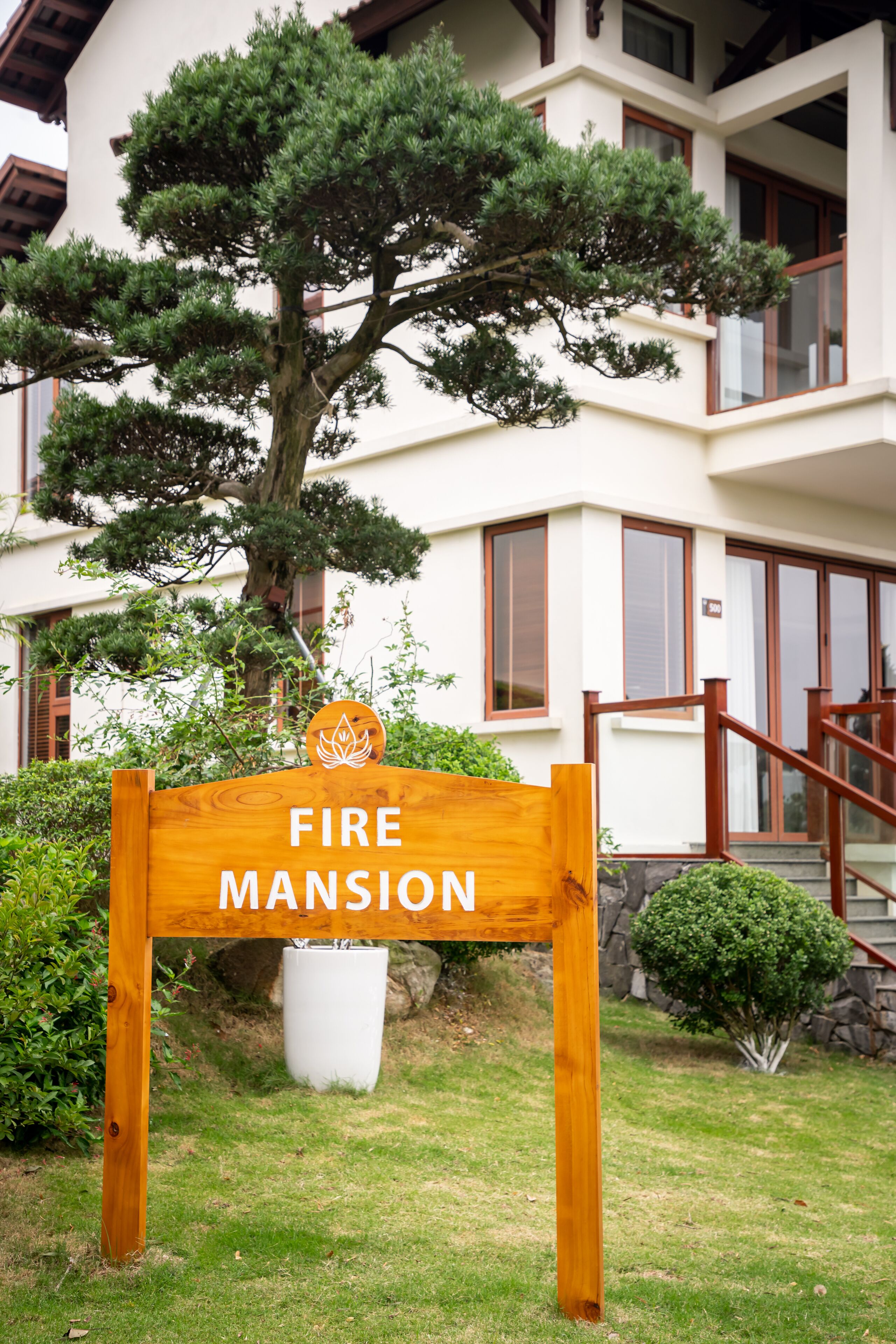 Uma placa de madeira com a inscrição 'FIRE MANSION', em frente a um edifício com uma árvore grande e um vaso branco.