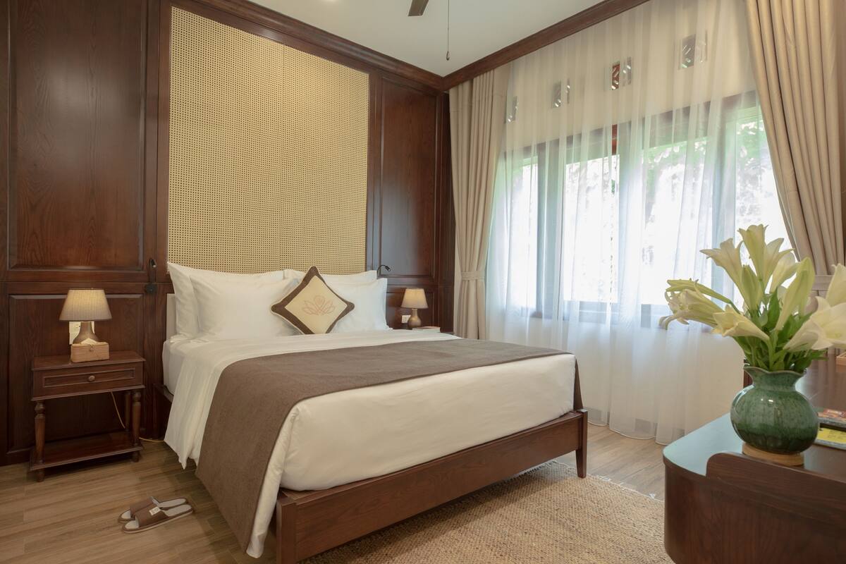 A neatly made bed with a brown bedspread, two bedside tables with lamps, a vase with flowers, and a window with sheer curtains.