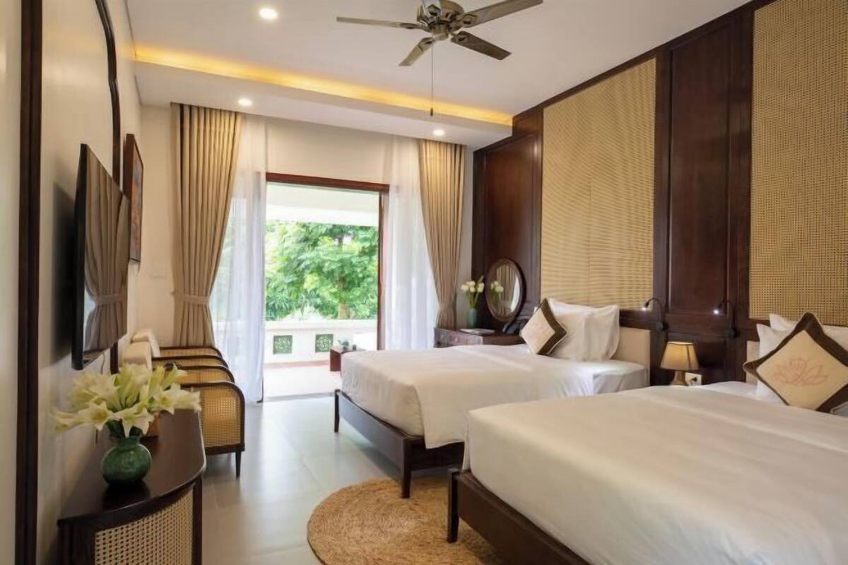 A hotel room with two beds, a ceiling fan, a large window with curtains, a bedside table with a vase of flowers, and a view of greenery outside.