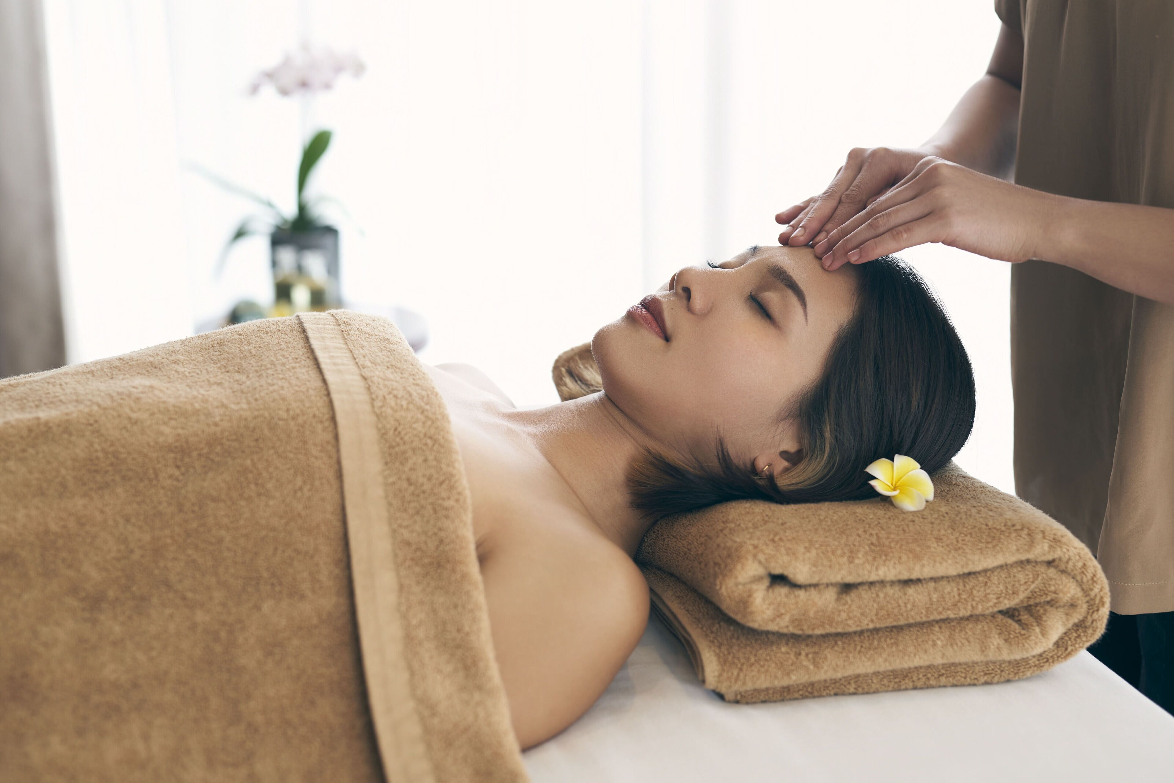 A person lying on a bed with a towel over their head, a flower in their hair, and a hand performing a massage.