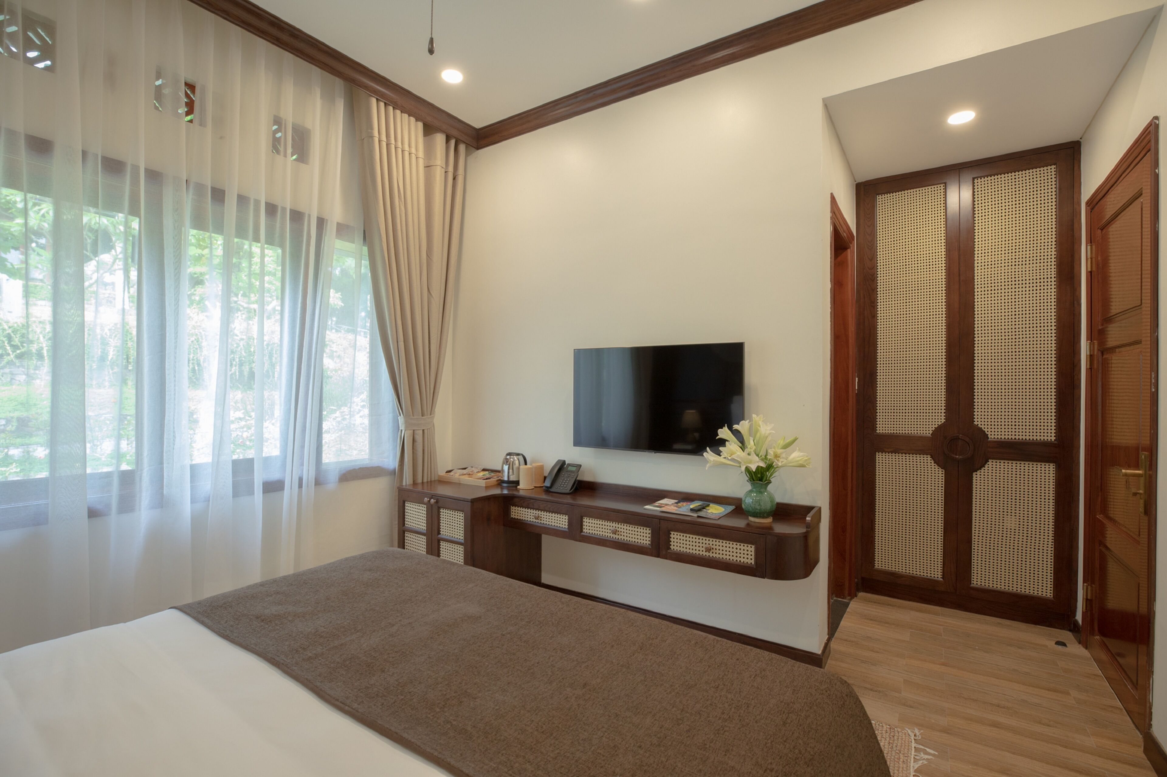 A hotel room with a bed, a TV mounted on the wall, a wooden console table with a vase of flowers, and a large wooden door with a decorative panel.
