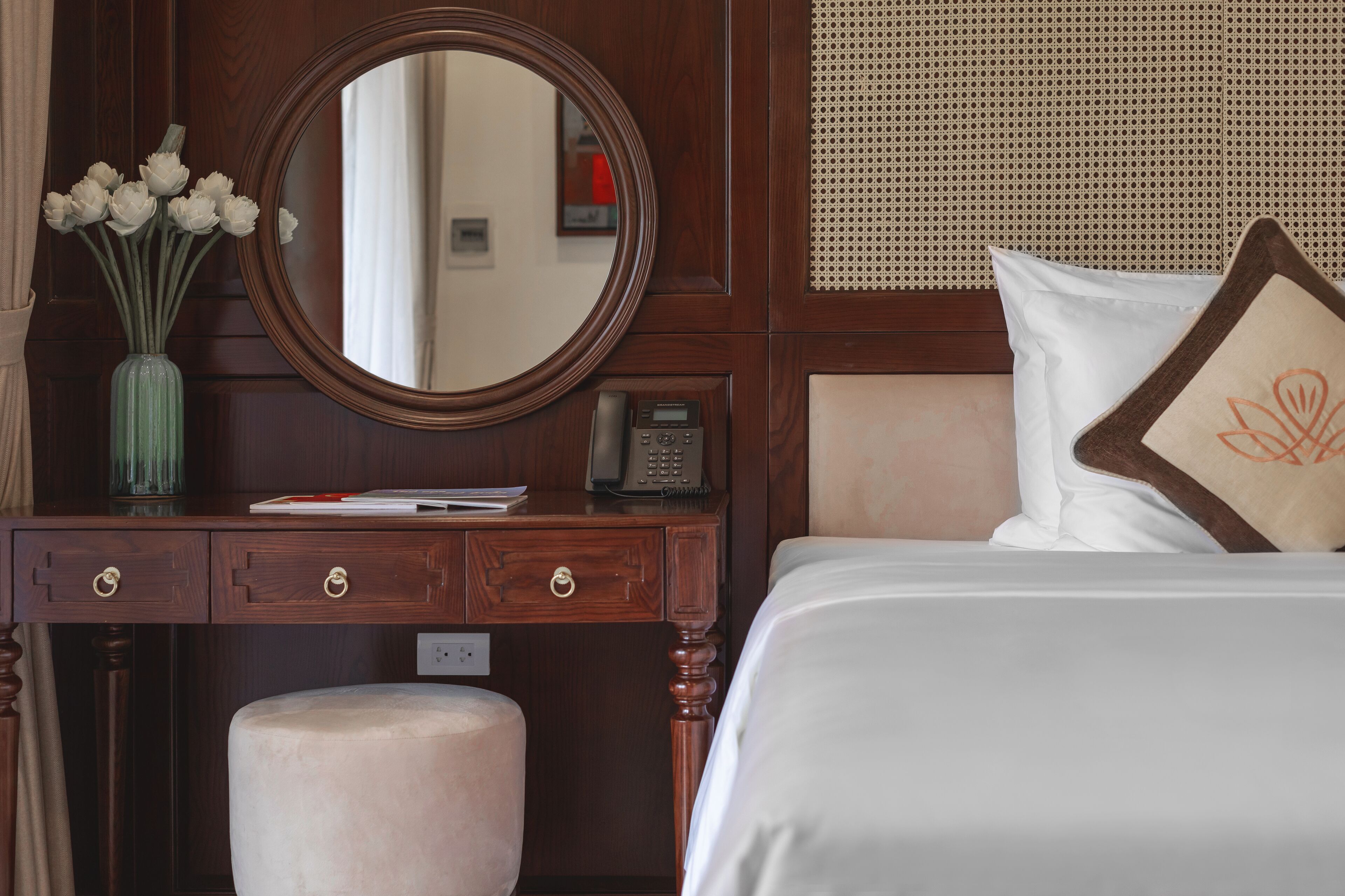 Una habitación de hotel con un aparador de madera, un espejo, un florero, un teléfono y una cama con un cojín.