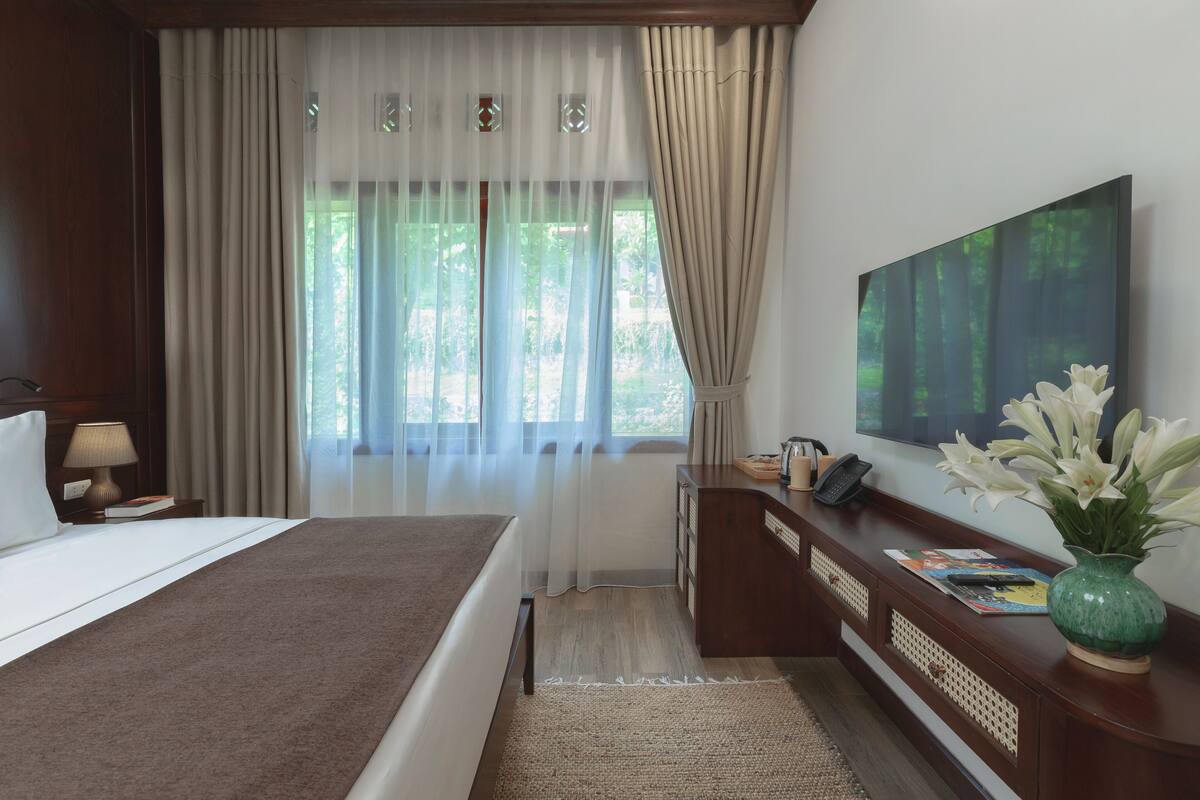 A hotel room with a large bed, a desk with a vase of flowers, a flat-screen TV, and a window with sheer curtains.