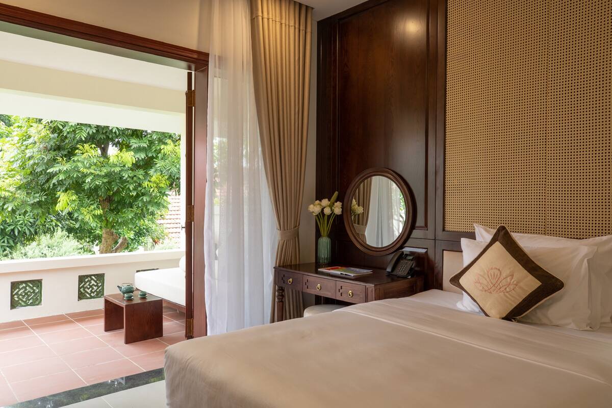 A hotel room with a bed, a vanity with a mirror, a small table with a vase of flowers, and a view of greenery outside.