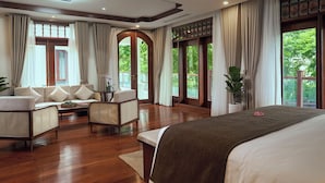 Um quarto espaçoso com uma cama grande, um sofá e uma mesa de jantar. Várias janelas oferecem luz natural e vista para o verde lá fora.