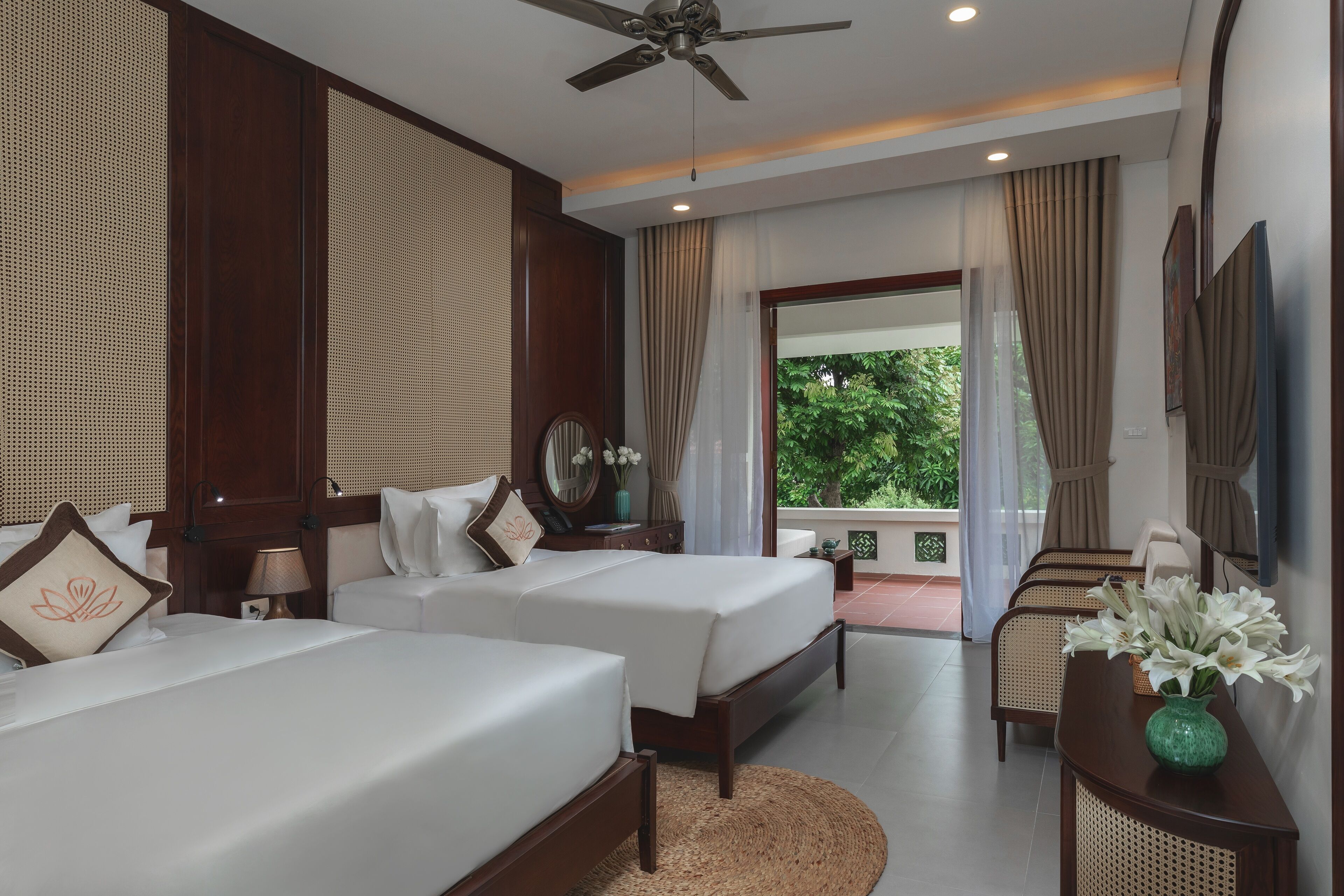 A hotel room with two beds, a ceiling fan, a large window with curtains, a small table with a vase, and a view of greenery outside.