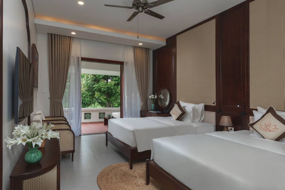 A hotel room with two beds, a desk, a chair, a ceiling fan, and a view of greenery outside.