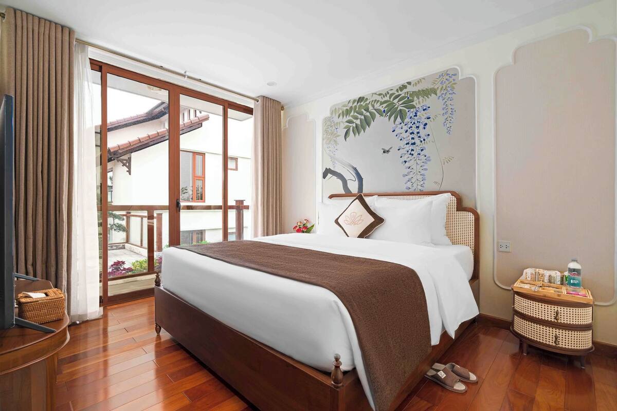 A hotel room with a large bed, a wooden floor, a sliding glass door to the balcony, and a wall-mounted TV.