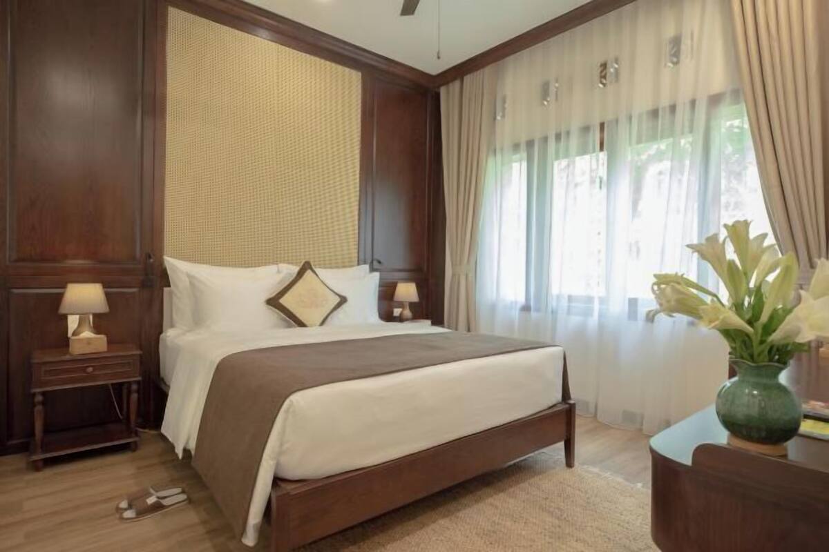 A hotel room with a large bed, wooden headboard, two bedside tables with lamps, a vase with flowers, and a view of greenery through the window.