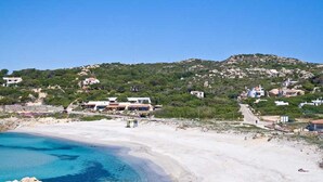 Une zone côtière avec une plage de sable, une eau d’un bleu limpide et quelques bâtiments dispersés sur une colline.