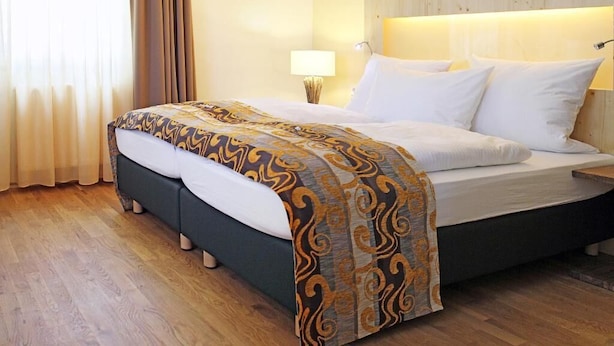 A hotel room with a bed, wooden flooring, a headboard, and a nightstand with a lamp. - Brauhotel & Gasthof GROSCH (Roedental)