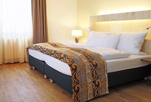 Quarto de hotel com cama, piso de madeira, cabeceira e mesa de cabeceira com abajur.