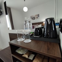 Habitación de hotel con una mesa de madera, dos vasos transparentes, dos botellas de agua, una tetera eléctrica negra y una cafetera.