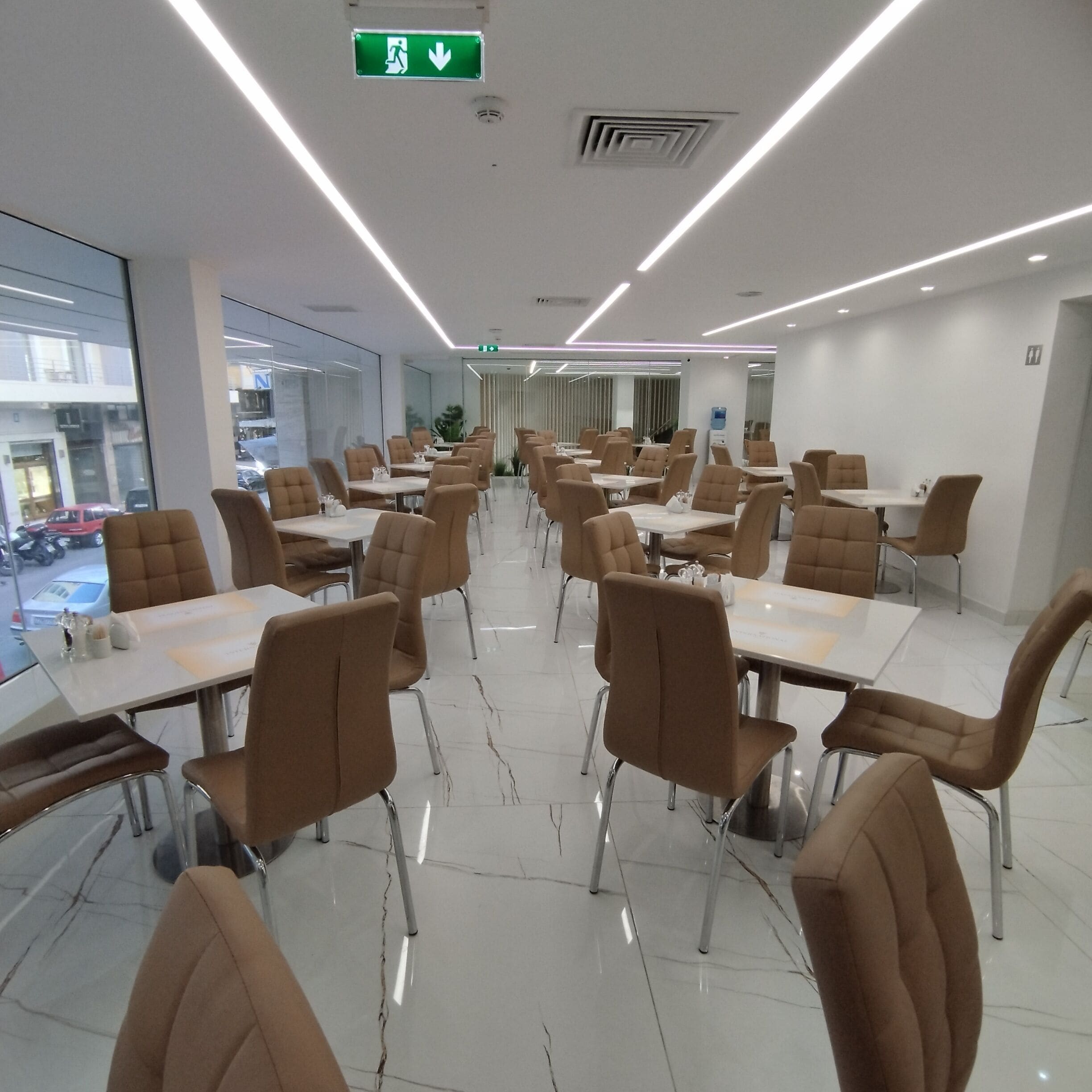 A modern dining area with tables and chairs, a visible exit sign, and a view of the street outside.