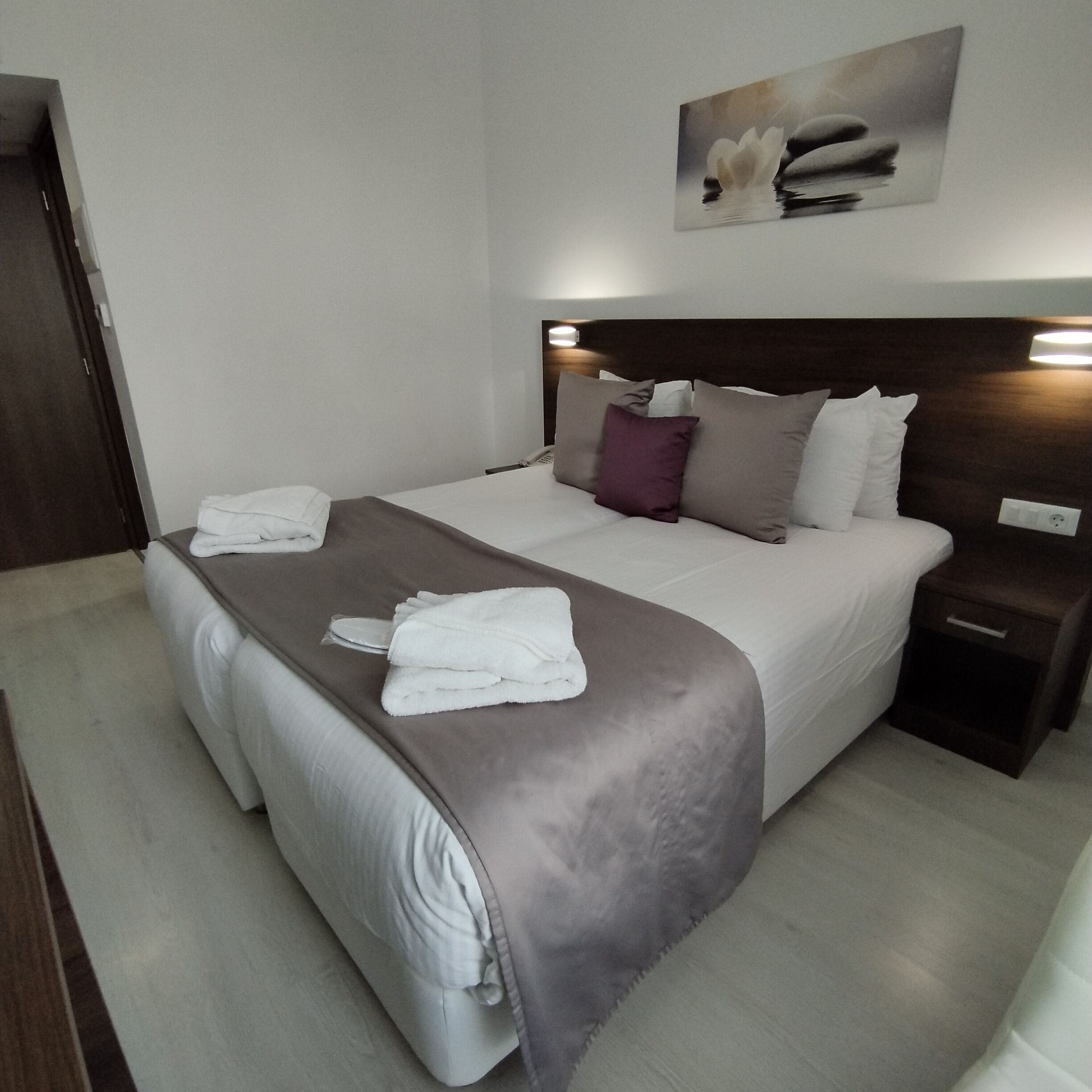 Een netjes opgemaakt bed met een grijs-witte dekbedovertrek, een bruine plaid en een witte handdoek die op het bed is gevouwen.