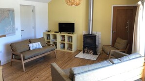 Een gezellige woonkamer met een houten vloer, een bank, een televisie op een dressoir, een boekenkast en een houtkachel.
