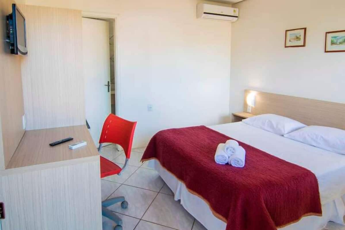 A hotel room with a bed, a desk, a chair, an air conditioning unit, and a TV.