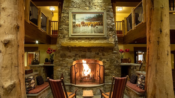 A stone fireplace with a framed painting above it, two rocking chairs, and a wooden bench in a cozy lobby area. - Ski-in Ski-out Luxurious 3 Bed 3 Bth + Free Ski Passes @ Teton Club (Jackson)