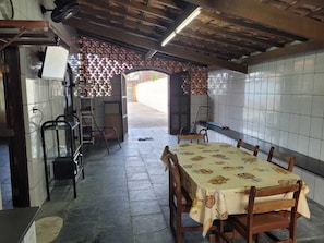A dining area with a table set for four, surrounded by chairs. There is a tiled wall on one side and a wooden ceiling. An open doorway leads to an outdoor area. - House with Pool in Itanhaém – Family and Pets (Itanhaém)