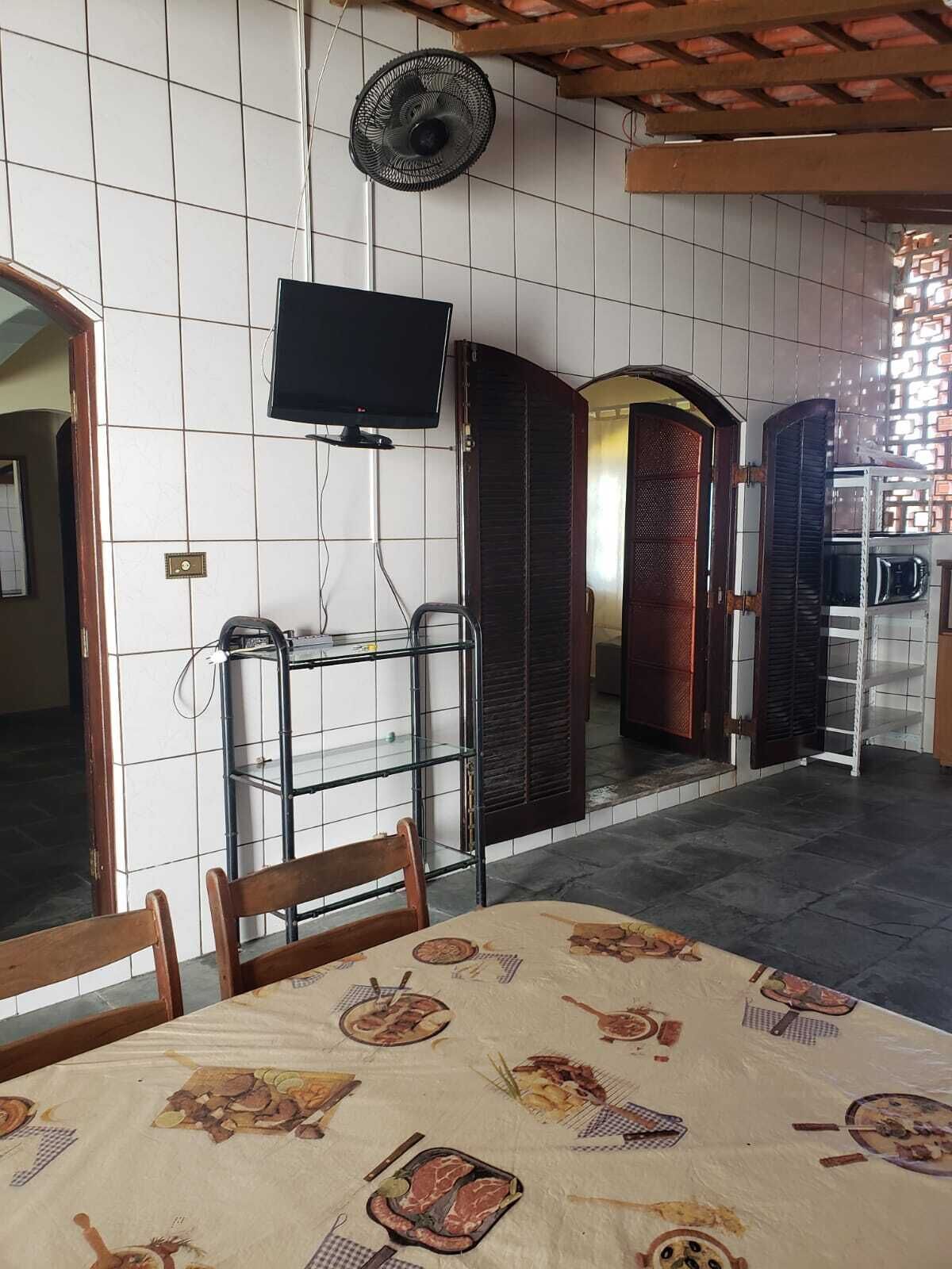 A room with a tiled wall, a television mounted on the wall, a fan, a table with a tablecloth, and a door with a mirror.
