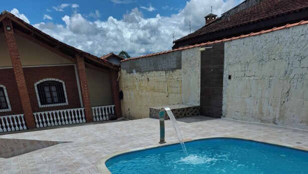 A swimming pool with a tiled deck, a water fountain, and a building with a tiled roof and a white wall in the background. - House with Pool in Itanhaém – Family and Pets (Itanhaém)