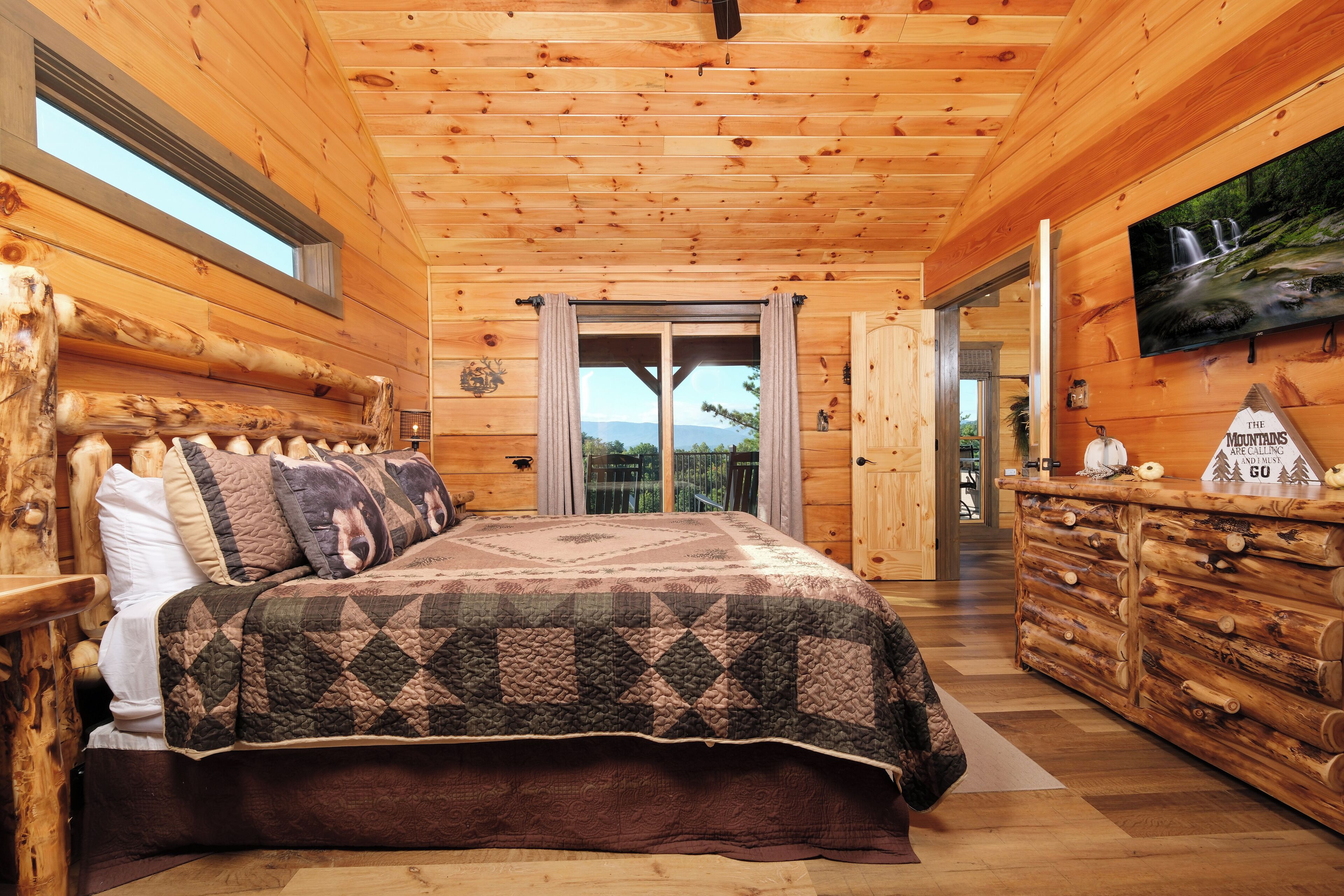 A log cabin bedroom with a large bed, wooden headboard, a dresser, and a TV mounted on the wall.