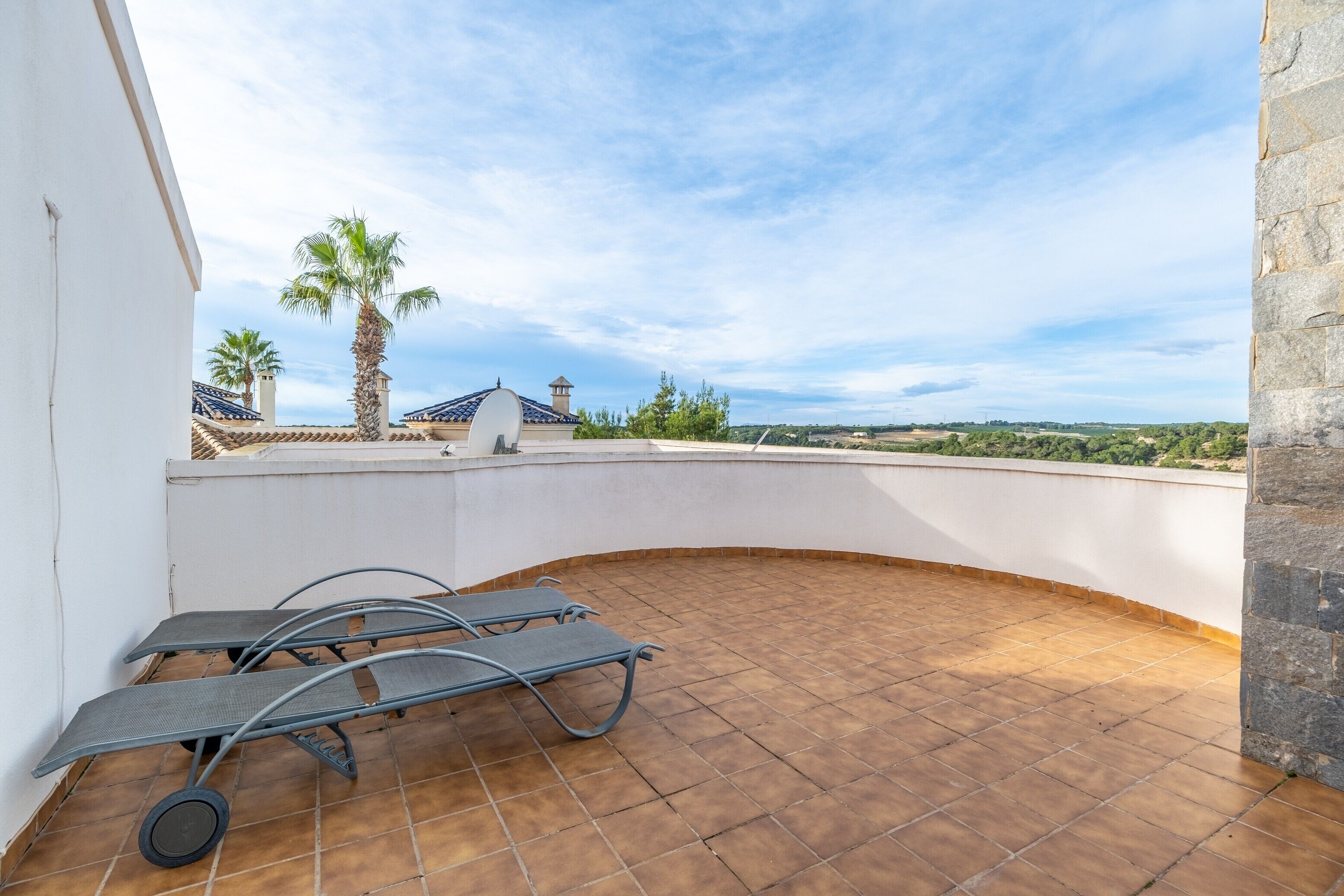 Terraza en la azotea con un muro blanco curvo, una tumbona de metal y vistas a palmeras y un paisaje lejano.