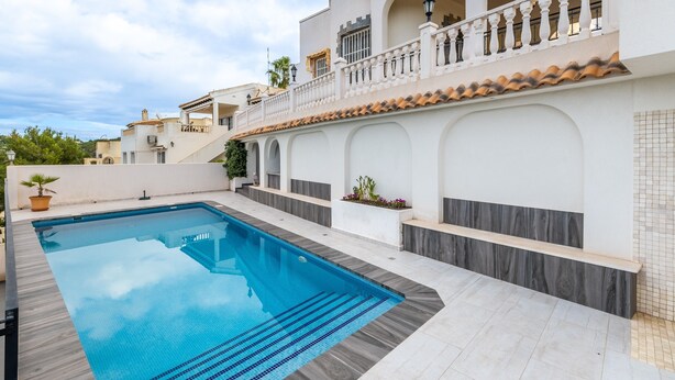 Una piscina con plataforma empaveda, un edificio blanco con arcos y un balcón con barandilla.