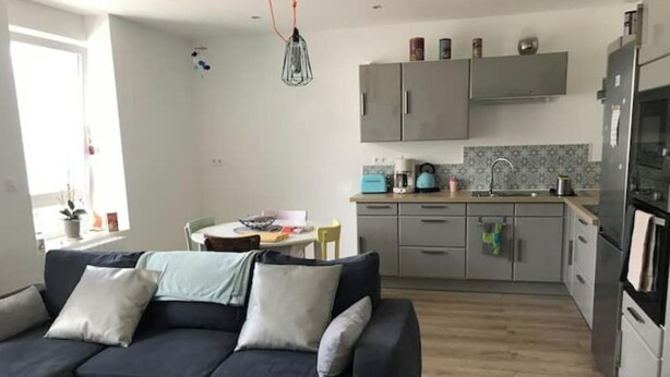 Una sala de estar moderna con un sofá oscuro, una mesa de centro redonda y un comedor con mesa y sillas. La cocina tiene armarios grises, un fregadero y una estufa.