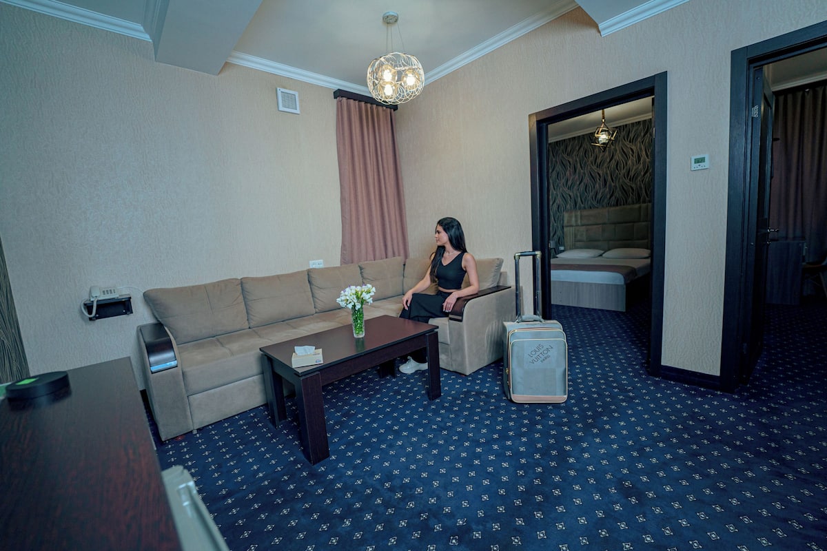Ein Hotelzimmer mit einer Couch, einem kleinen Tisch mit einer Vase, einem Koffer und einem Bett mit Kopfteil.
