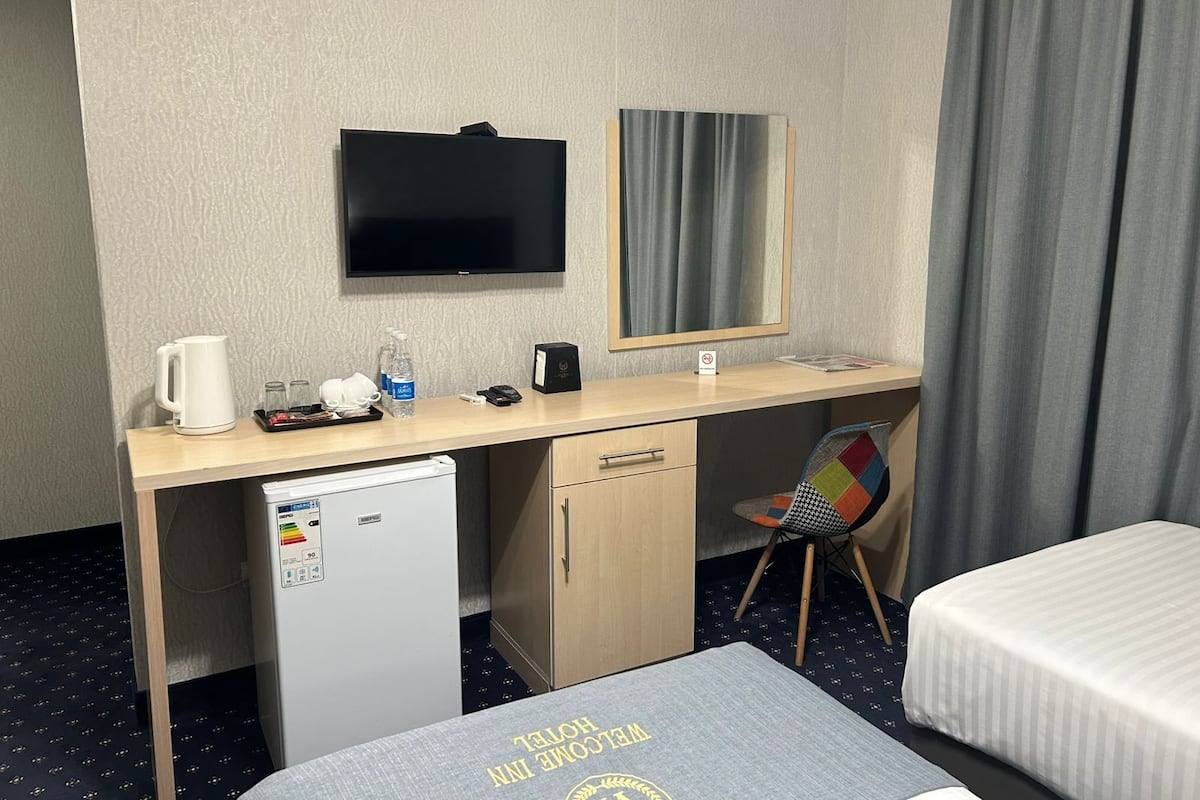 Ein Hotelzimmer mit einem Bett, einem Schreibtisch mit Fernseher, einem Kühlschrank, einem Sessel und einem fenster mit Vorhängen.