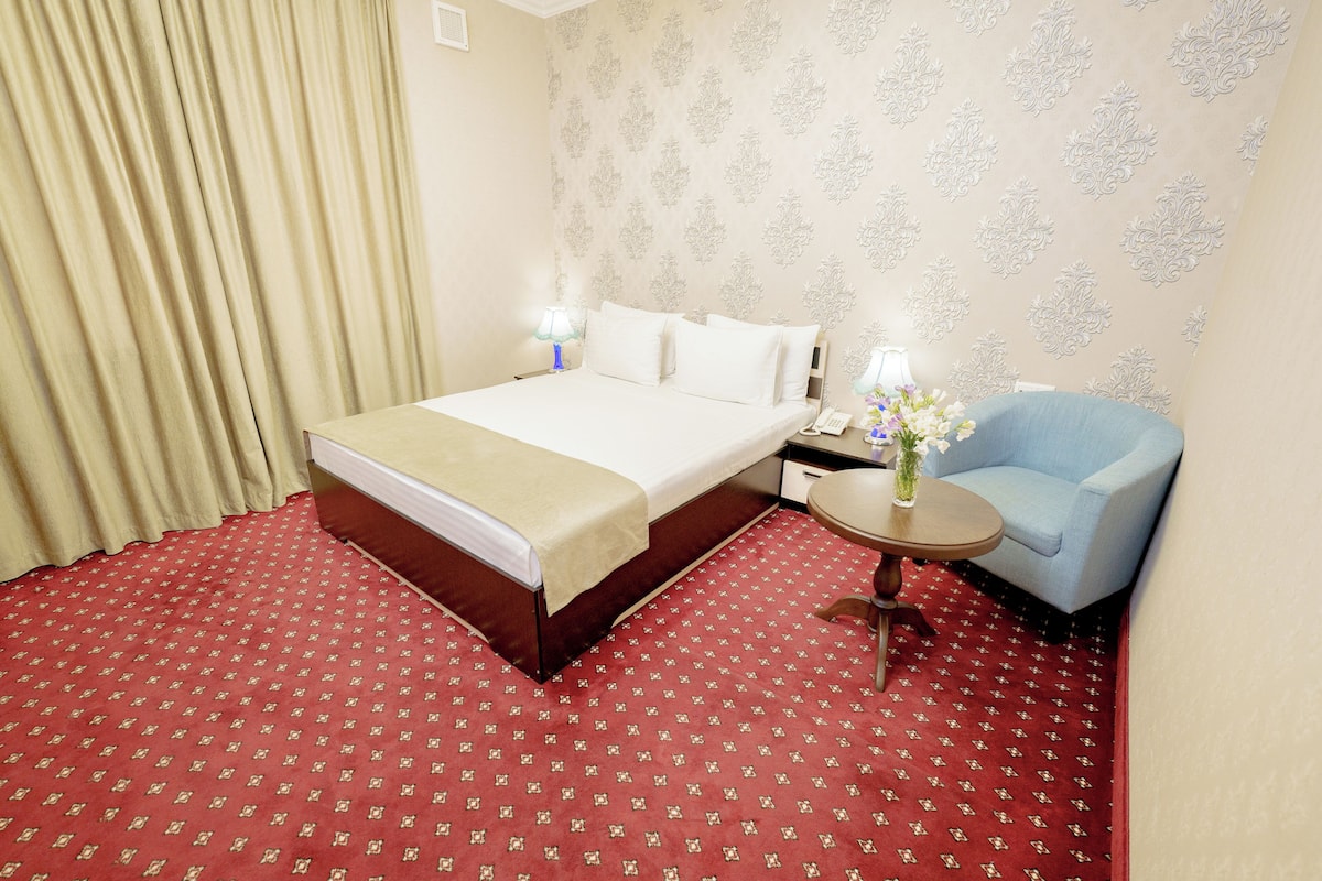Ein Hotelzimmer mit einem Bett, einem Sessel, einem kleinen Tisch mit einer Vase und einem gemusterten Teppich.