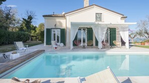 A poolside area with a covered seating area, a pool, and a house with green shutters.