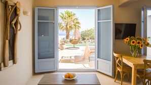 Porta deslizante aberta que dá para um alpendre com uma palmeira, uma cadeira de vime e uma mesa com uma tigela de frutas.